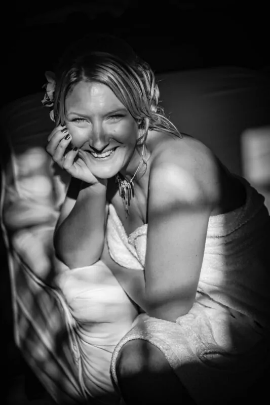 A joyful bride smiles broadly while resting her chin on her hand in a candid black and white getting ready portrait.