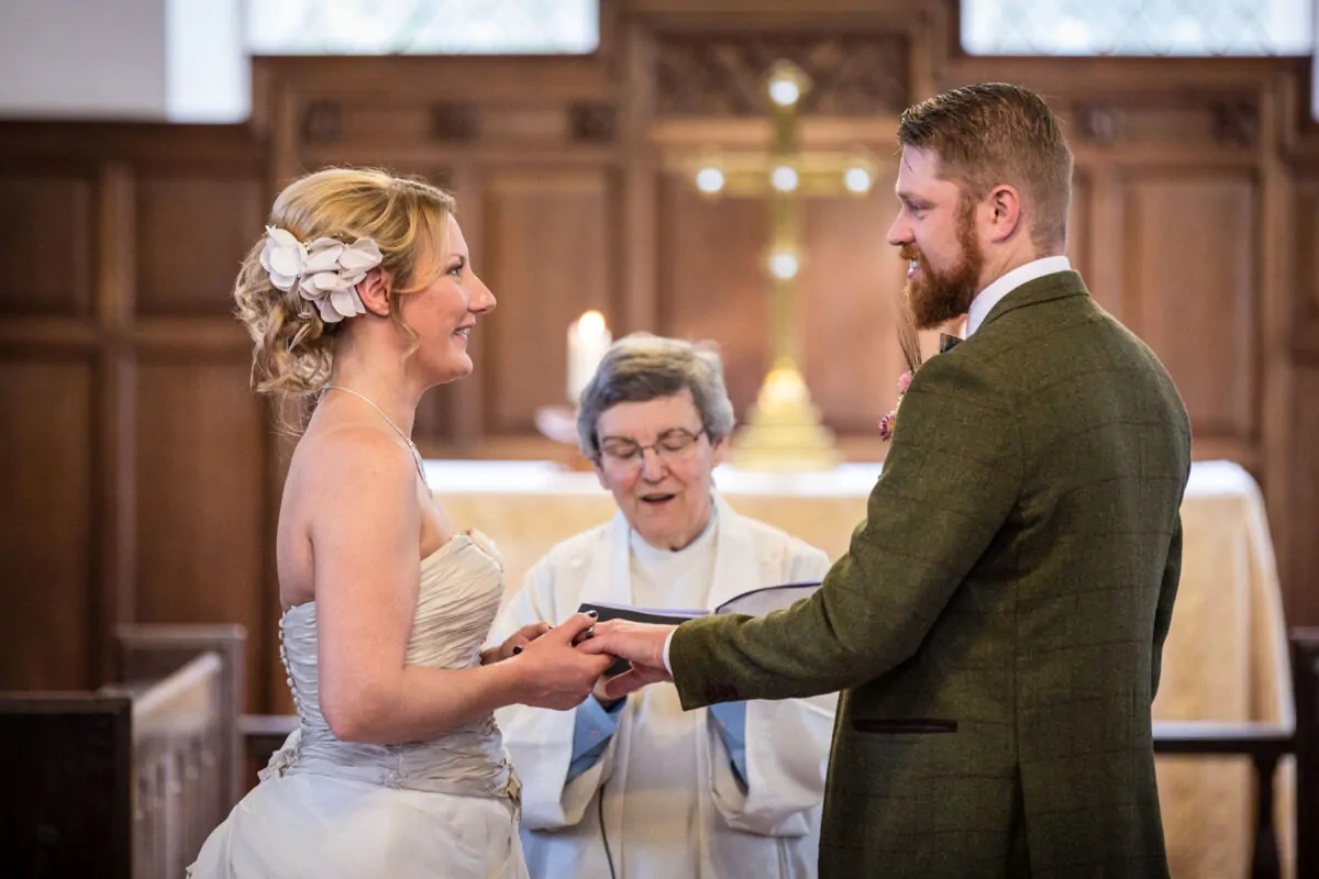 A bride and groom exchange rings during a church wedding ceremony, officiated by a celebrant standing between them.