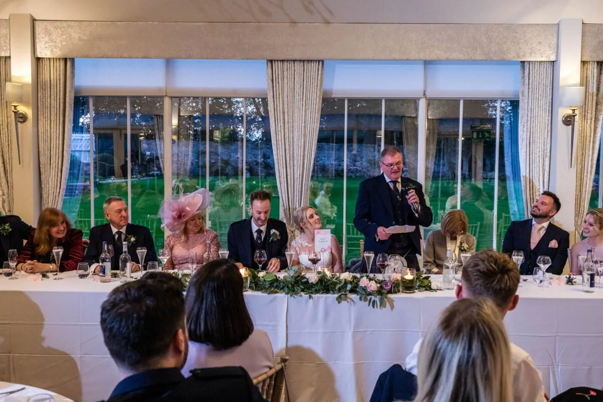A father of the bride delivers a speech at the top table during a wedding reception, while guests seated below look on.