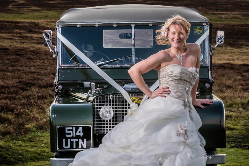 A smiling bride in a voluminous strapless gown poses beside a vintage green Land Rover against a dramatic moorland and stormy sky backdrop.