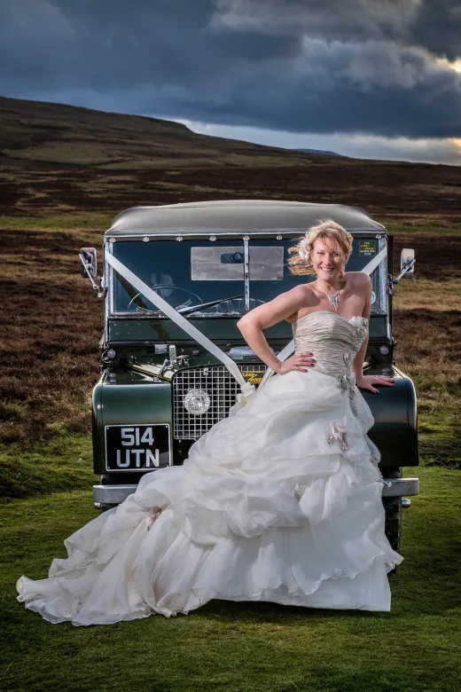 A smiling bride in a voluminous strapless gown poses beside a vintage green Land Rover against a dramatic moorland and stormy sky backdrop.