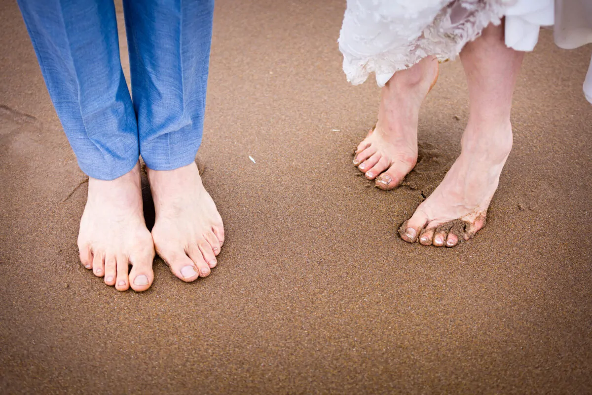 A close-up shot capturing the barefoot feet of two newlyweds standing on a wet sandy beach, with blue trouser cuffs on the left and a white lace dress hem on the right.