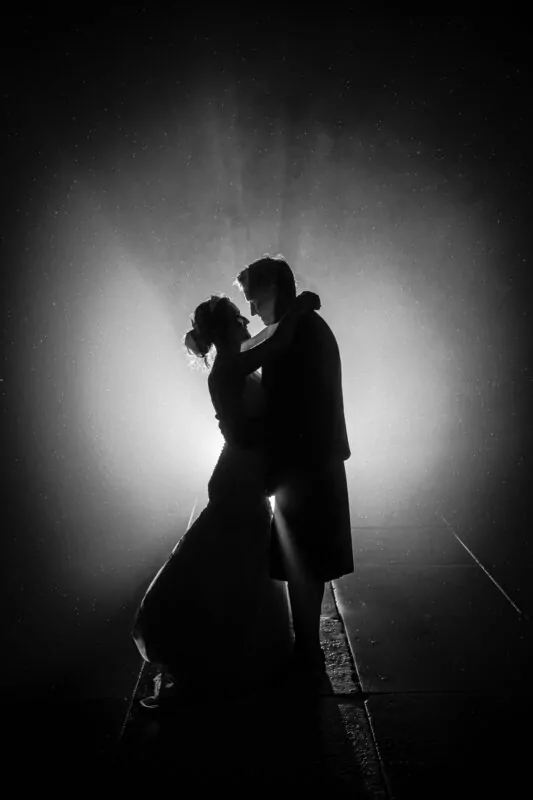 A dramatic black and white silhouette of a bride and groom embracing under a starry night sky with atmospheric backlighting.