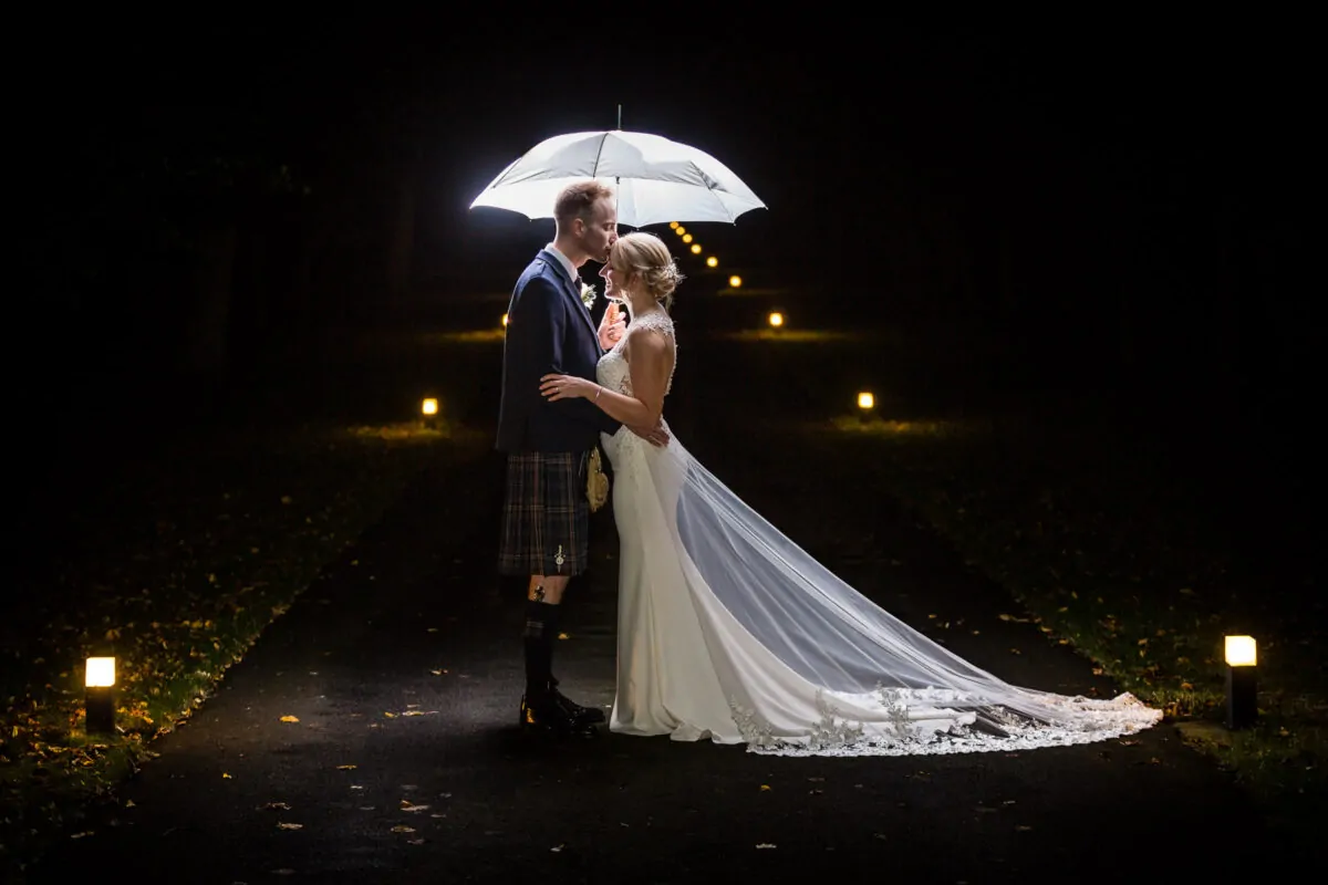 A newlywed couple shares a tender forehead kiss under an illuminated umbrella on a dark pathway lined with glowing lights at night.