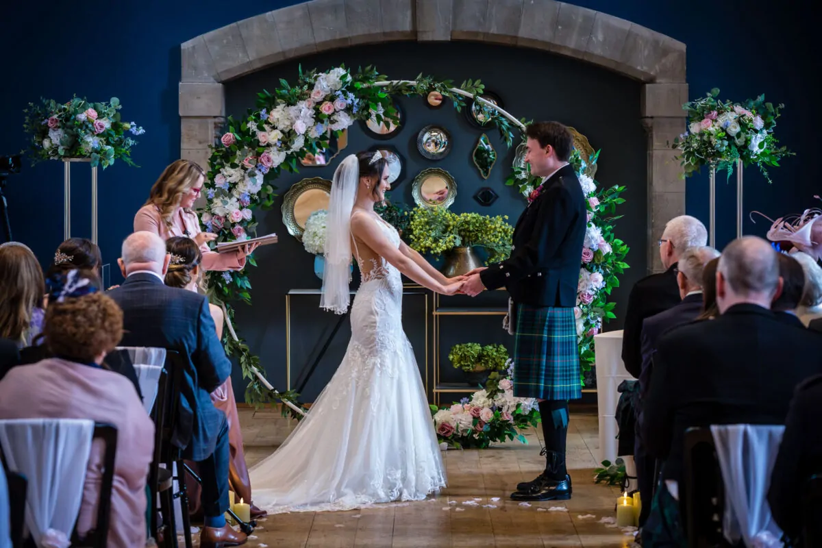 A bride in a lace gown and veil holds hands with a groom in a kartan kilt during their ceremony in front of a large floral arch.