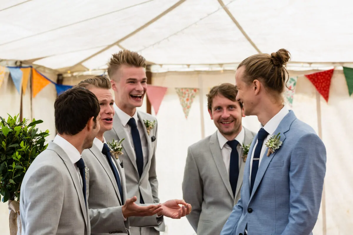 A group of groomsmen in grey and blue suits share a joyful moment chatting beneath a marquee decorated with colourful bunting.