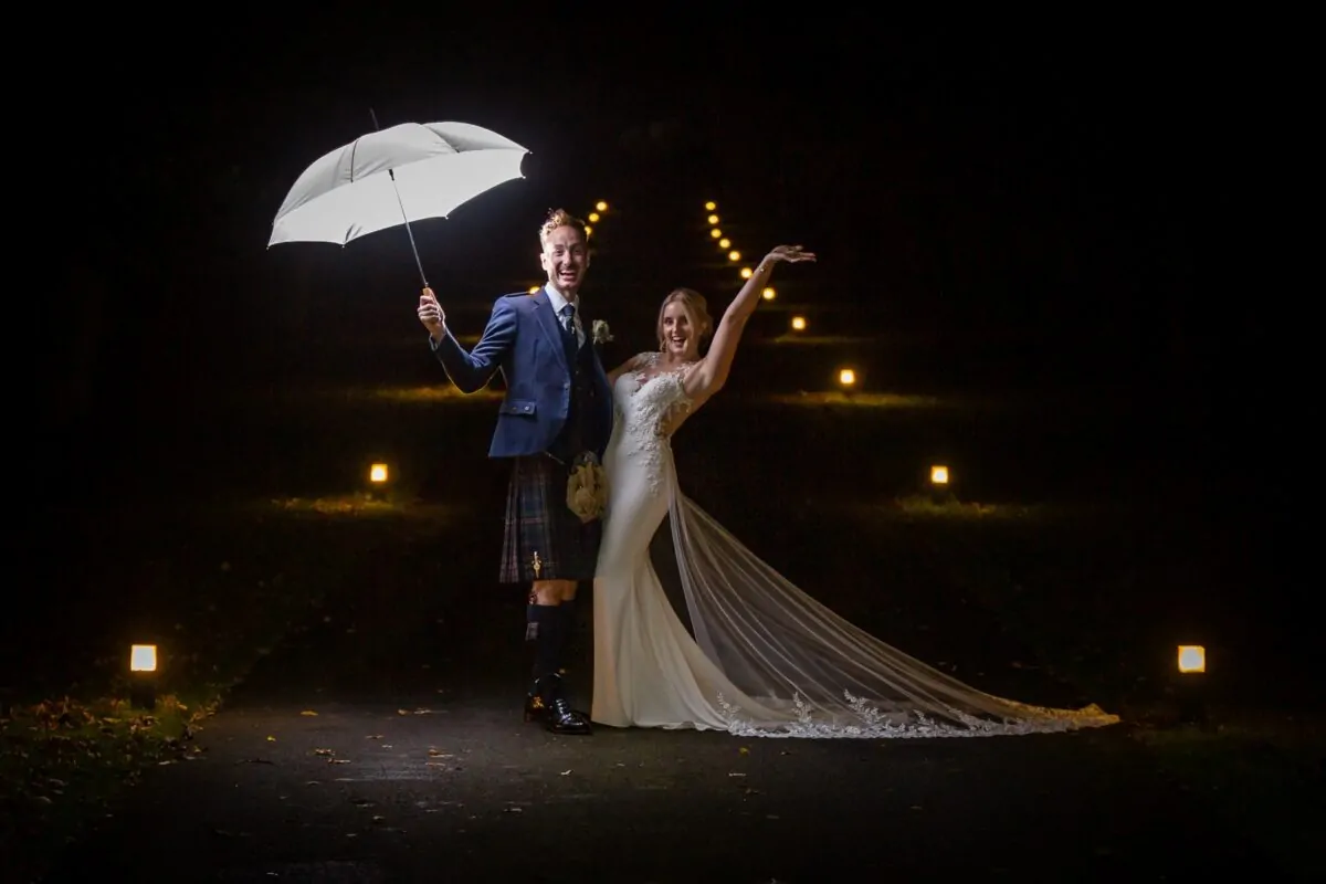 A newlywed couple poses joyfully on a path at night, illuminated under a glowing white umbrella with distant lights lining the background.
