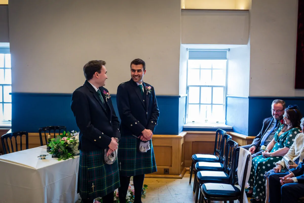 Two grooms in traditional tartan kilts smile at each other during their ceremony inside a bright room with seated guests.