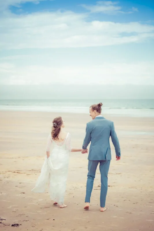 Newlyweds walk hand in hand barefoot along a sandy beach under a blue sky, with the bride wearing a lace gown and the groom in a blue suit.