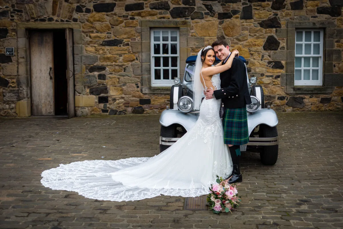 A newlywed couple poses in front of a vintage car parked outside a stone barn, with the bride wearing a dramatic lace gown featuring an extended train.