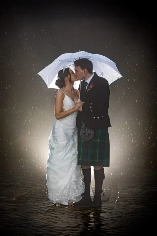 A bride and groom share a romantic kiss under a clear umbrella on a wet cobbled courtyard at night during a rainy wedding portrait.