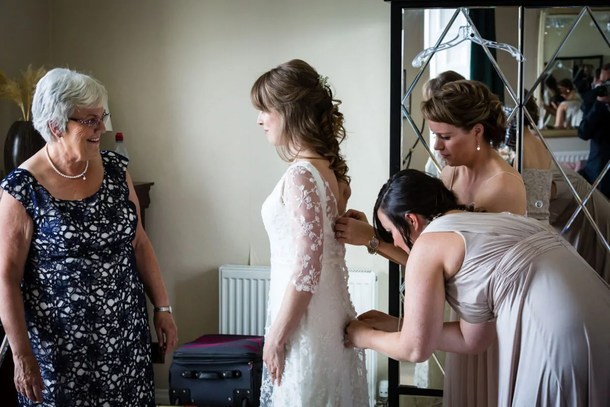 An older woman smiling warmly as bridesmaids help the bride button up her intricate lace wedding dress during morning preparations.