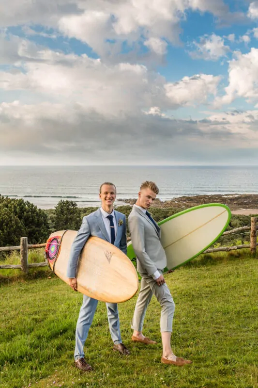 Two grooms in light grey suits happily pose with surfboards on a grassy clifftop overlooking the sea under a dramatic cloudy sky.