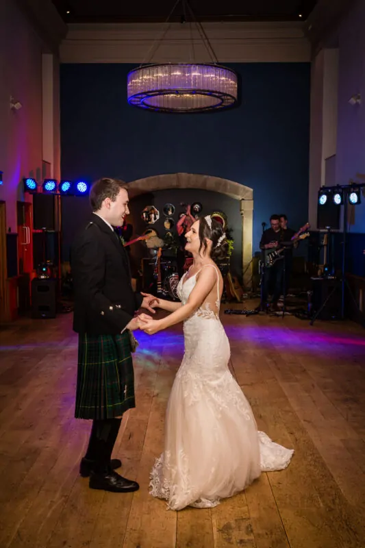 A bride in a lace wedding dress and a groom in a traditional tartan kilt share a joyful first dance on a wooden floor under colourful stage lights.
