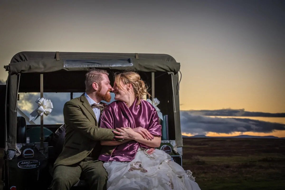 A newlywed couple shares a tender moment sitting in the back of a vintage Land Rover during a dramatic sunset.