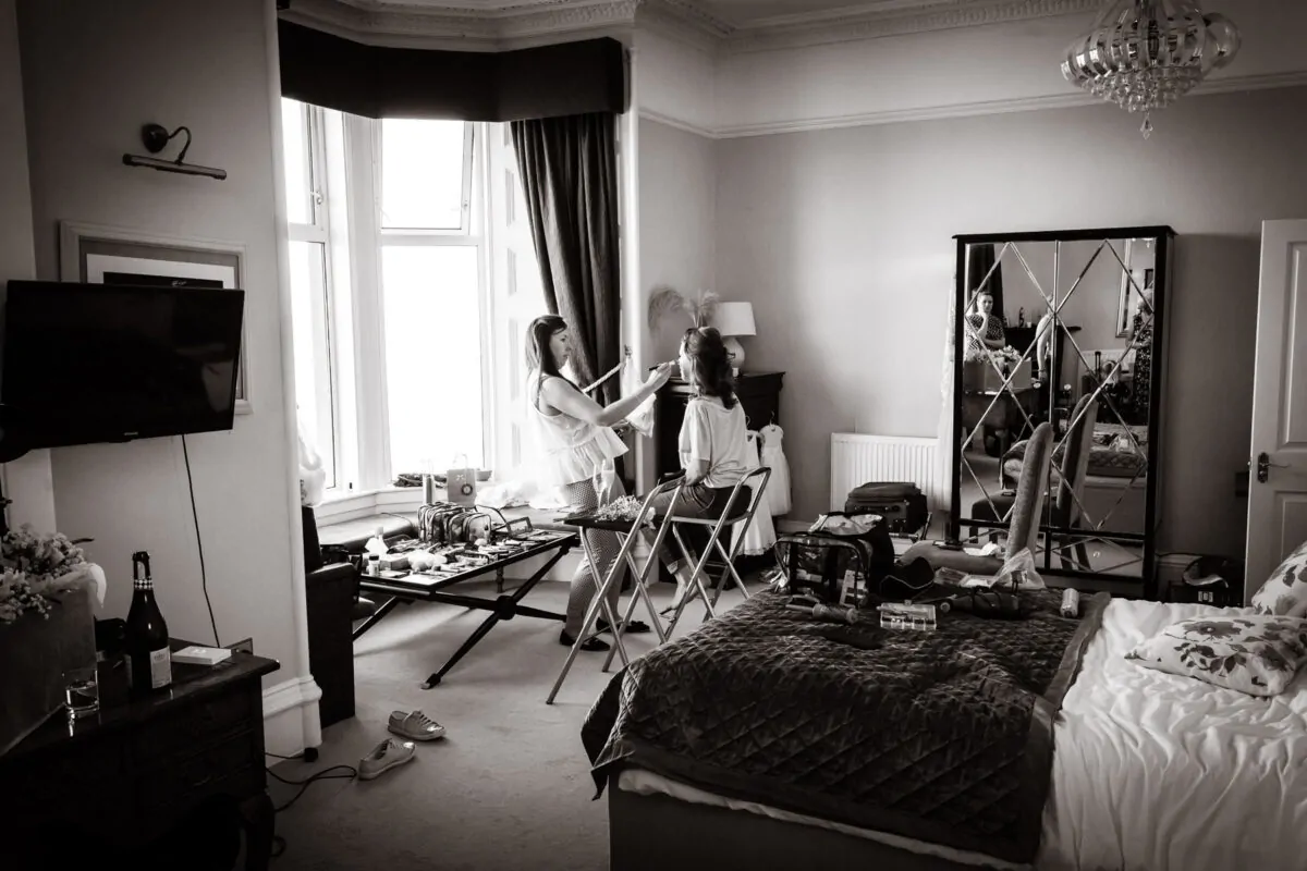 A bride sits calmly by a large bay window while a makeup artist meticulously applies finishing touches in a spacious, light-filled hotel bedroom.