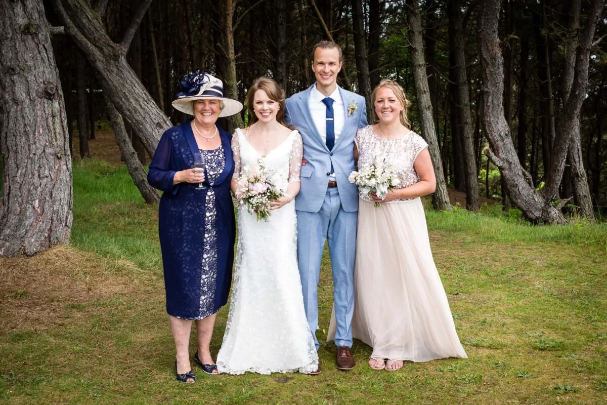 A newlywed couple stand outdoors in a woodland setting with the groom in a light blue suit and the bride in a white lace gown, flanked by a woman in a blue dress with a fascinator and a bridesmaid in a sequinned top and long skirt holding bouquets.