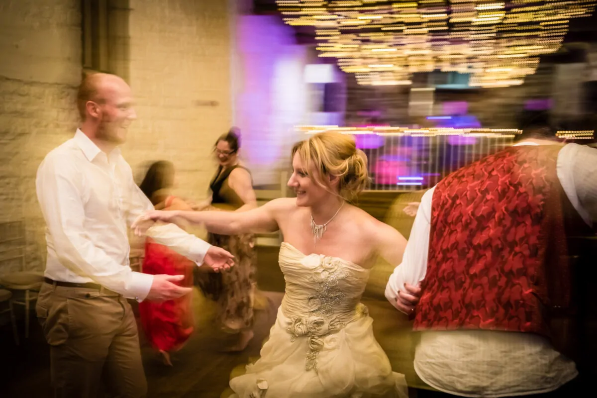 A bride in a strapless floral appliqué gown dances joyfully on a lively, motion-blurred dance floor beneath a canopy of twinkling lights.