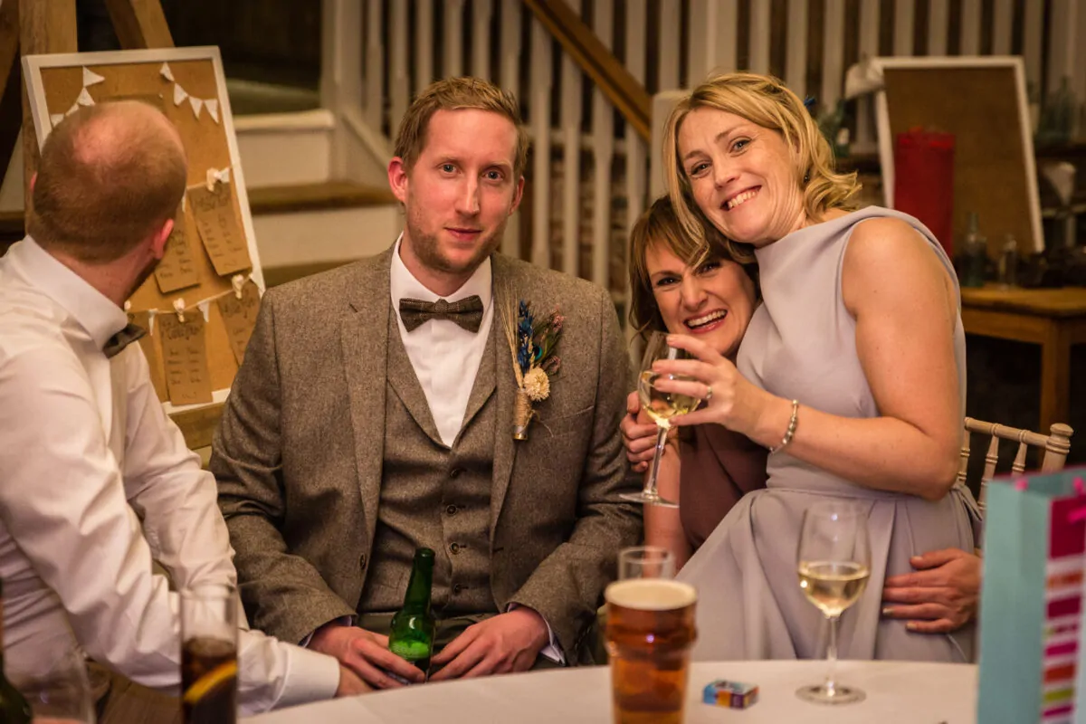 Guests smile warmly around a wedding breakfast table featuring a tweed-suited groom, a glass of wine, and a seating plan display in the background.