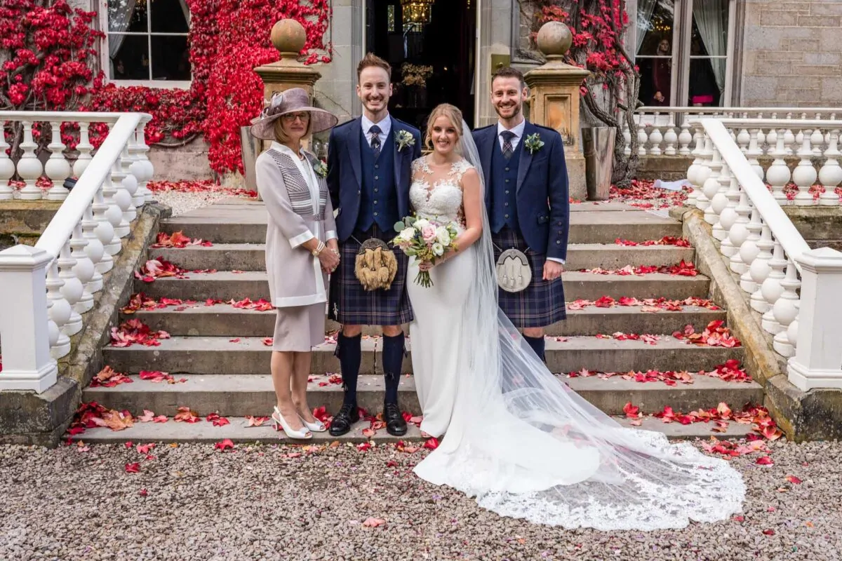 A bridal party including a bride in a lace gown, two groomsmen in tartan kilts, and a guest wearing a fascinator pose together on stone steps framed by an ivy-covered building and autumn leaves.