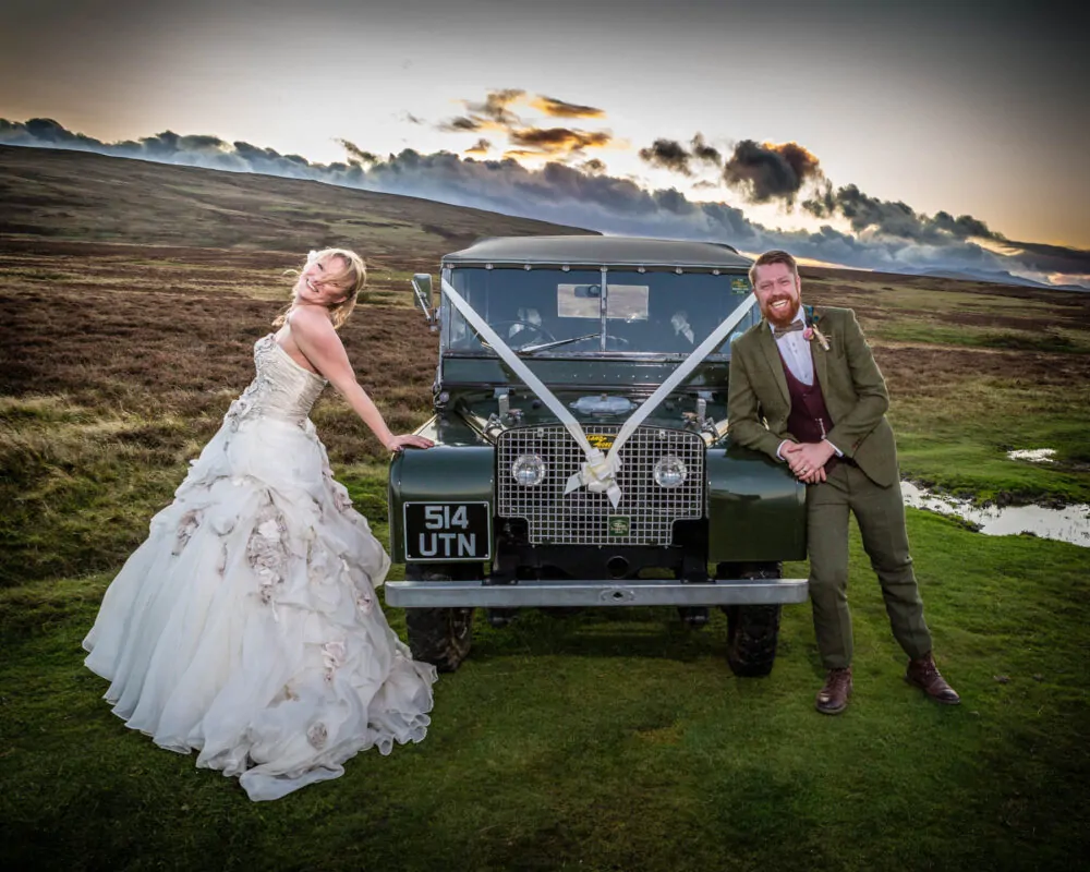 A joyful bride and groom lean against a classic green Land Rover decorated with ribbons amidst a dramatic moorland landscape at sunset.