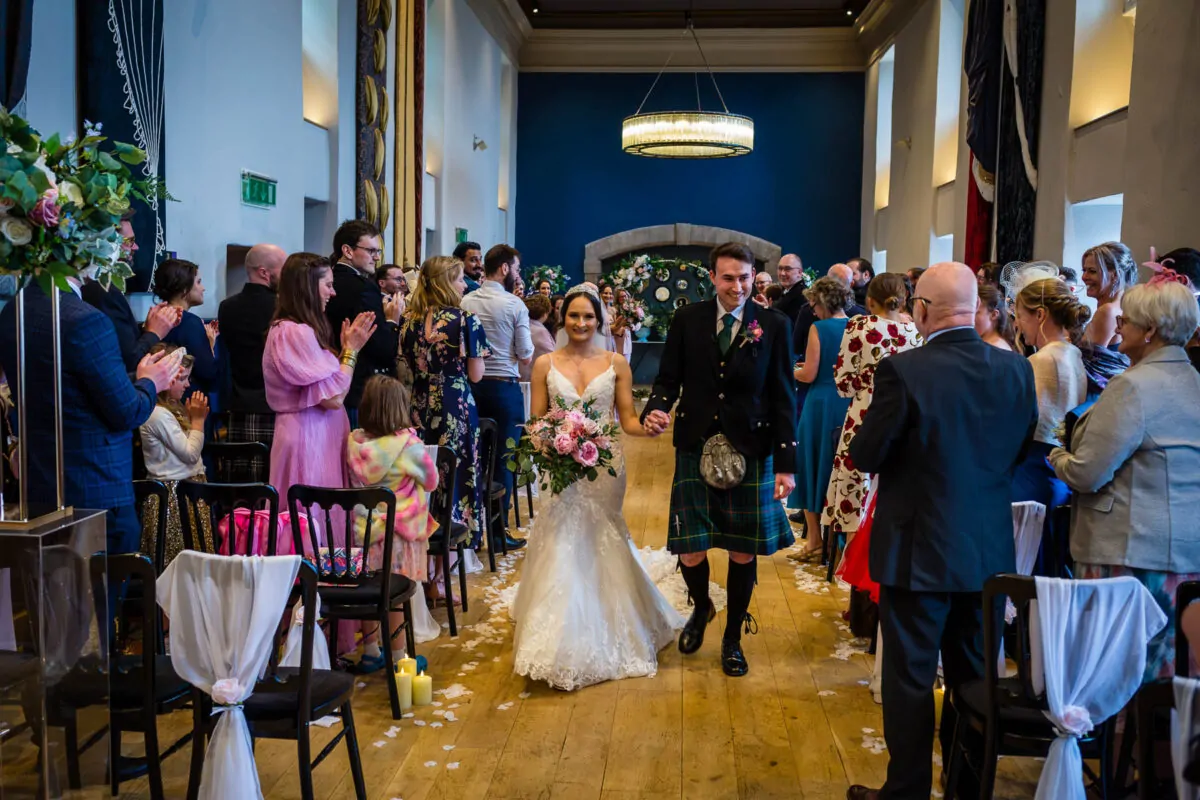 Newlyweds happily walk hand-in-hand down the aisle as guests applaud and celebrate during the recessional.
