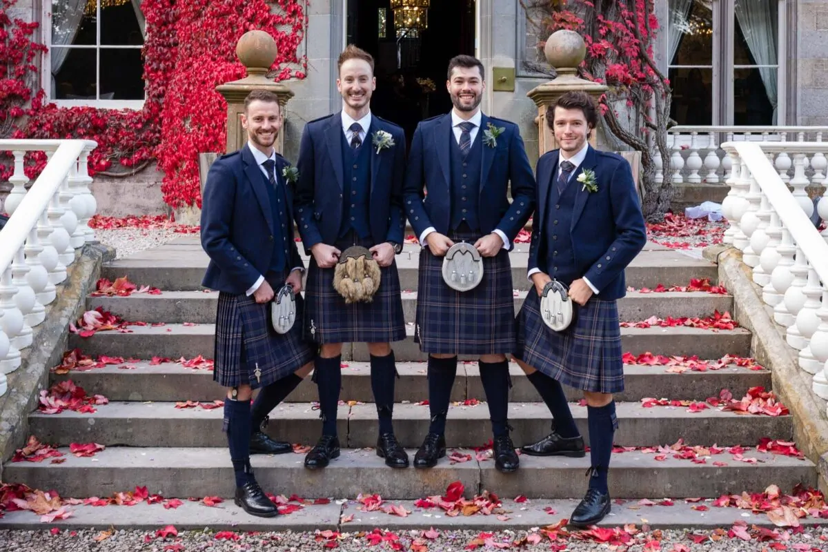 Four smiling groomsmen in traditional Scottish kilts, tweed jackets, and sporrans pose together on stone steps outside a stately venue surrounded by autumn leaves.