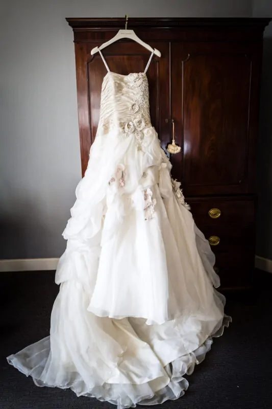 An ivory strapless wedding dress featuring a ruched bodice, crystal embellishments, and cascading floral applique hangs elegantly against a dark wooden wardrobe.