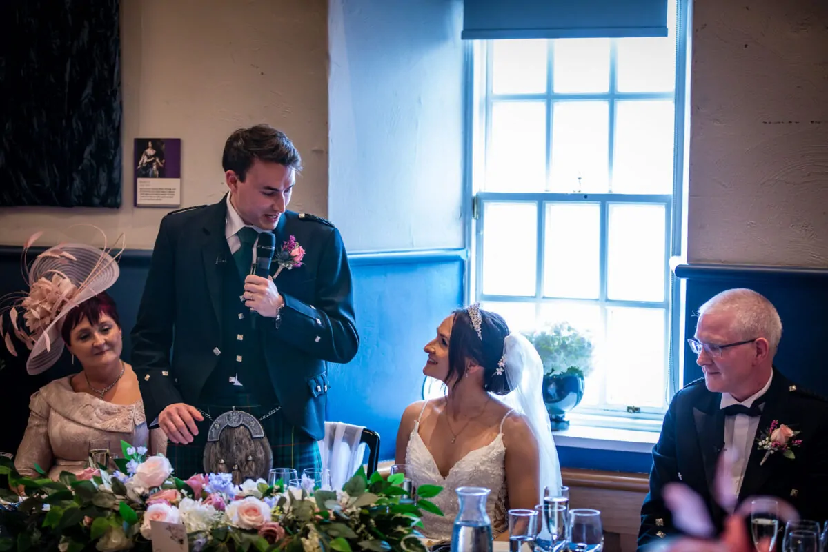 A groom in a traditional kilt gives a speech at the top table while his smiling bride looks up at him next to a sunlit window.