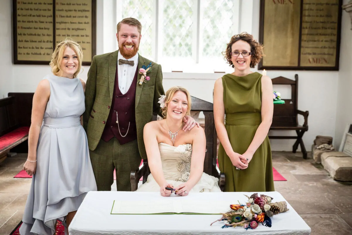 The bride smiles while signing the register at a church wedding, accompanied by the groom in a tweed suit and two bridesmaids wearing contrasting dresses.