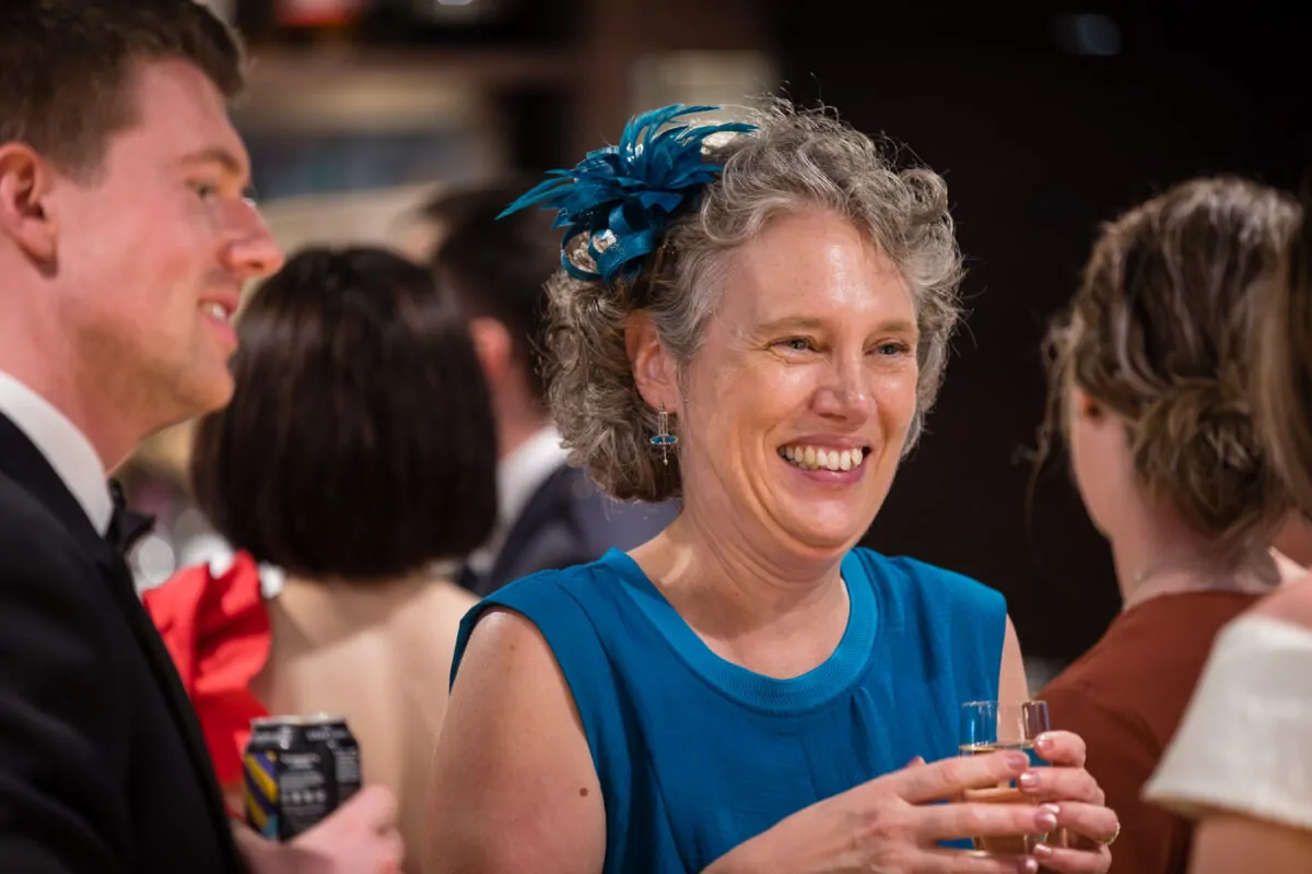 A smiling guest wearing a blue dress and a teal fascinator laughs while mingling at a wedding reception.