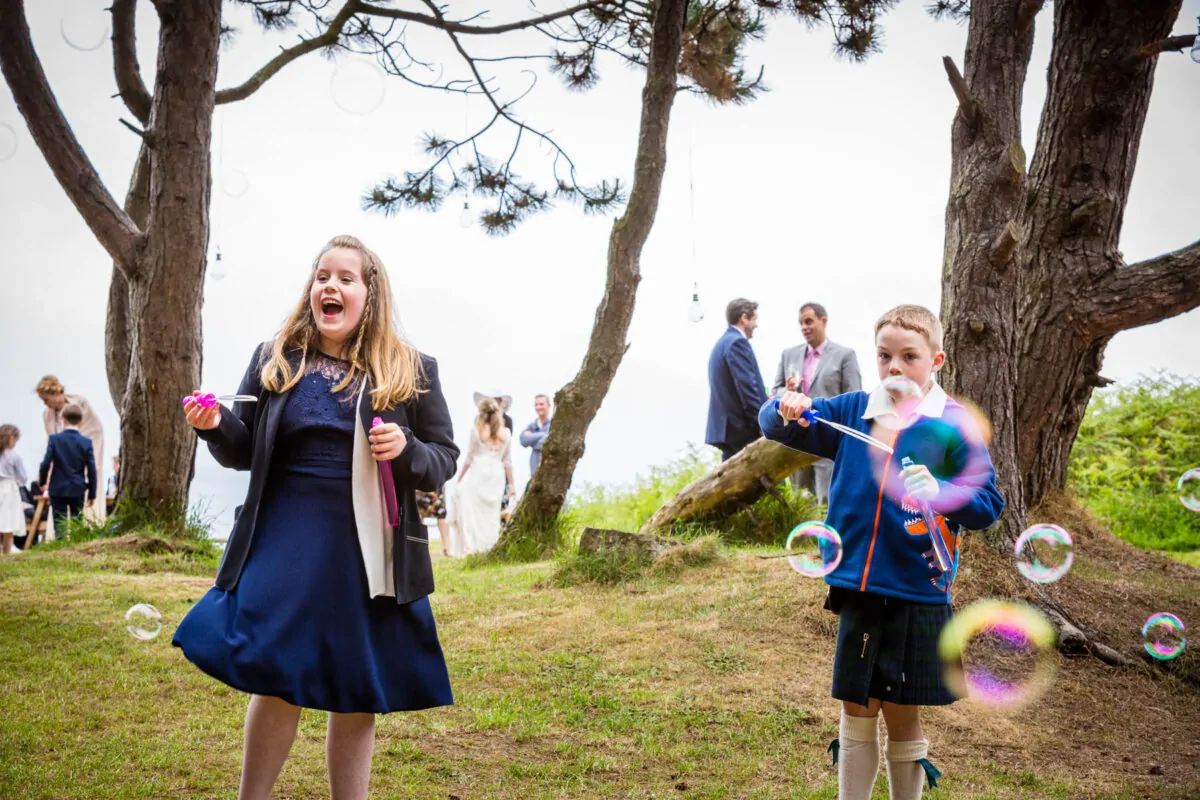 A young flower girl and a page boy in a kilt joyfully blow bubbles on a grassy lawn during an outdoor wedding celebration.