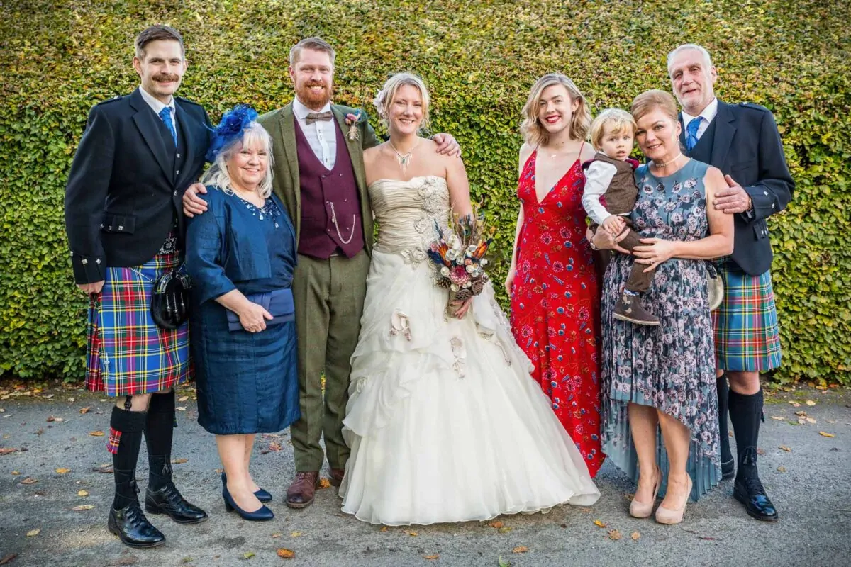 A happy wedding party, including the bride and groom in traditional highland dress and tweed, pose together outdoors in front of a dense green hedge.