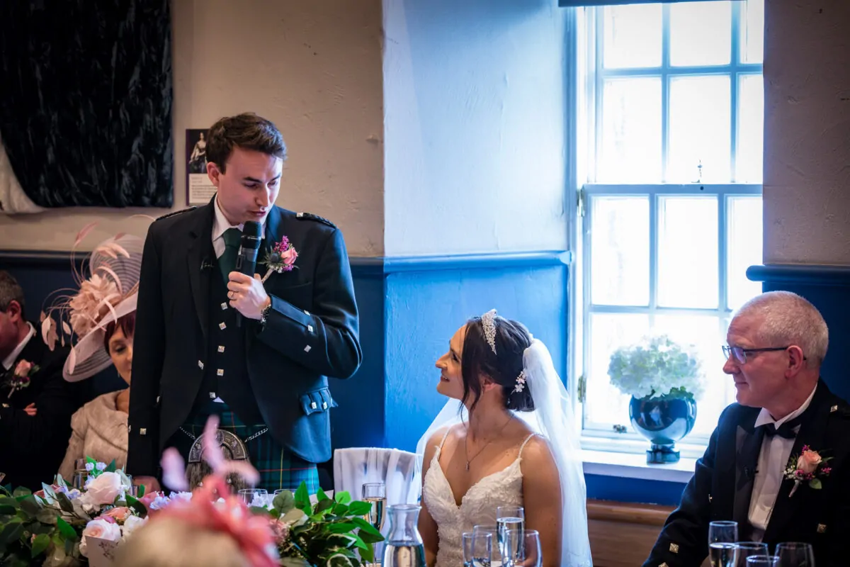 A groom wearing a traditional highland kilt gives a speech into a microphone at the reception table while looking down affectionately at his smiling bride, who wears a v-neck gown and tiara.