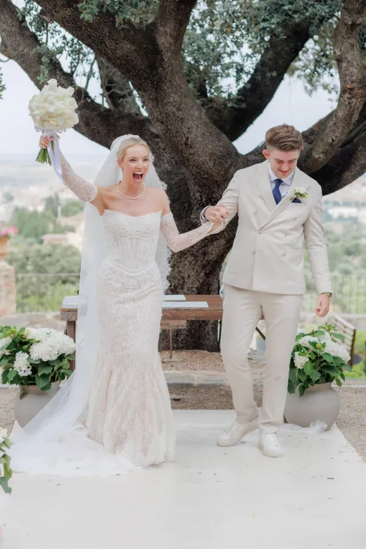 A newlywed couple joyfully walks away from their outdoor ceremony hand in hand, with the bride raising her bouquet in celebration beneath a grand oak tree.