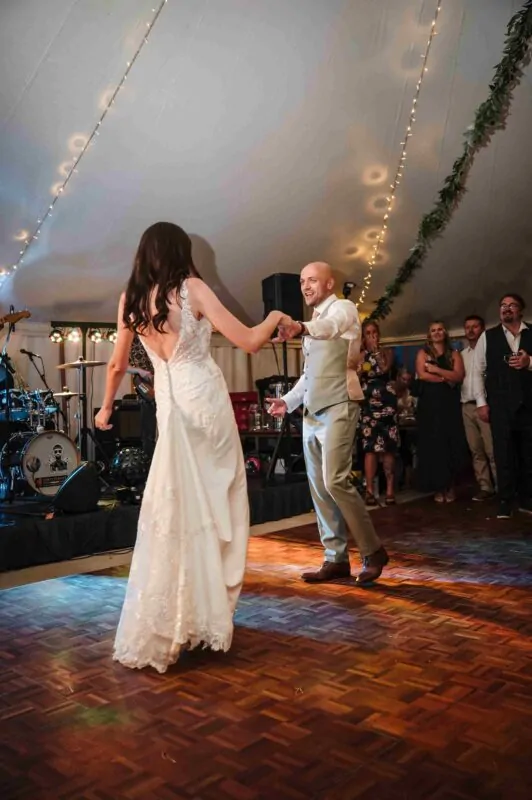 The newly married couple share their first dance together on a wooden dance floor inside a marquee illuminated by string lights.