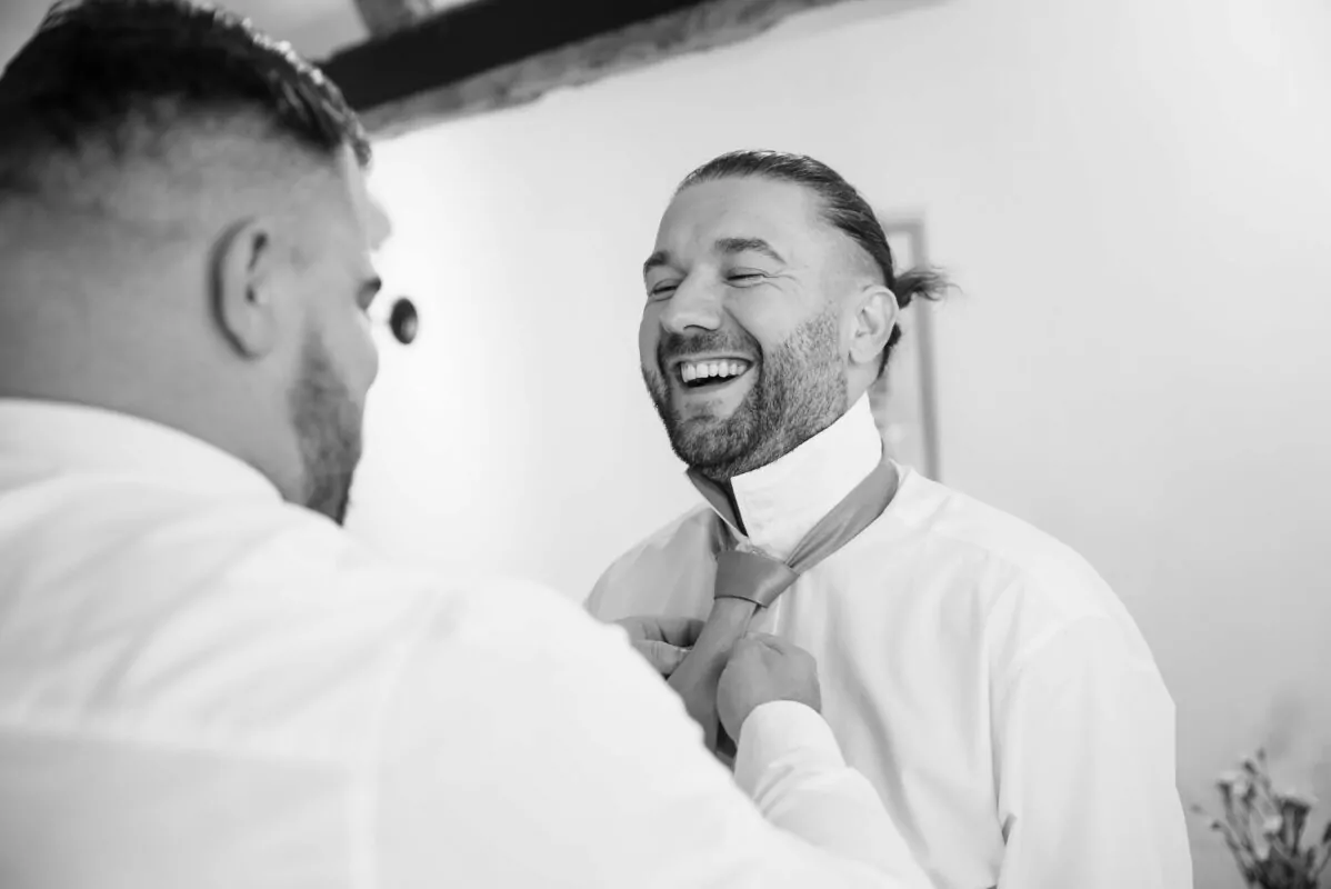 A joyful groom laughs aloud as his partner helps adjust his tie during morning preparations in a candid black and white portrait.