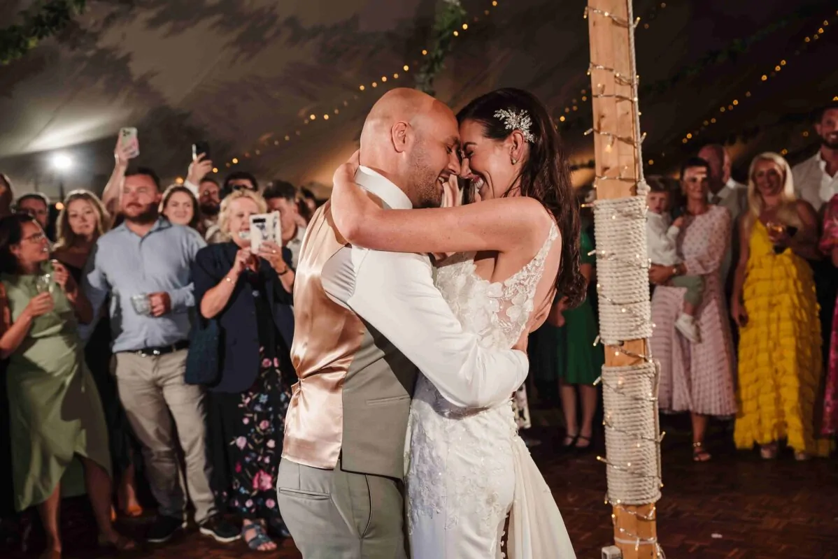 The bride and groom share a joyful laugh during their first dance inside a marquee illuminated by string lights, surrounded by cheering wedding guests.