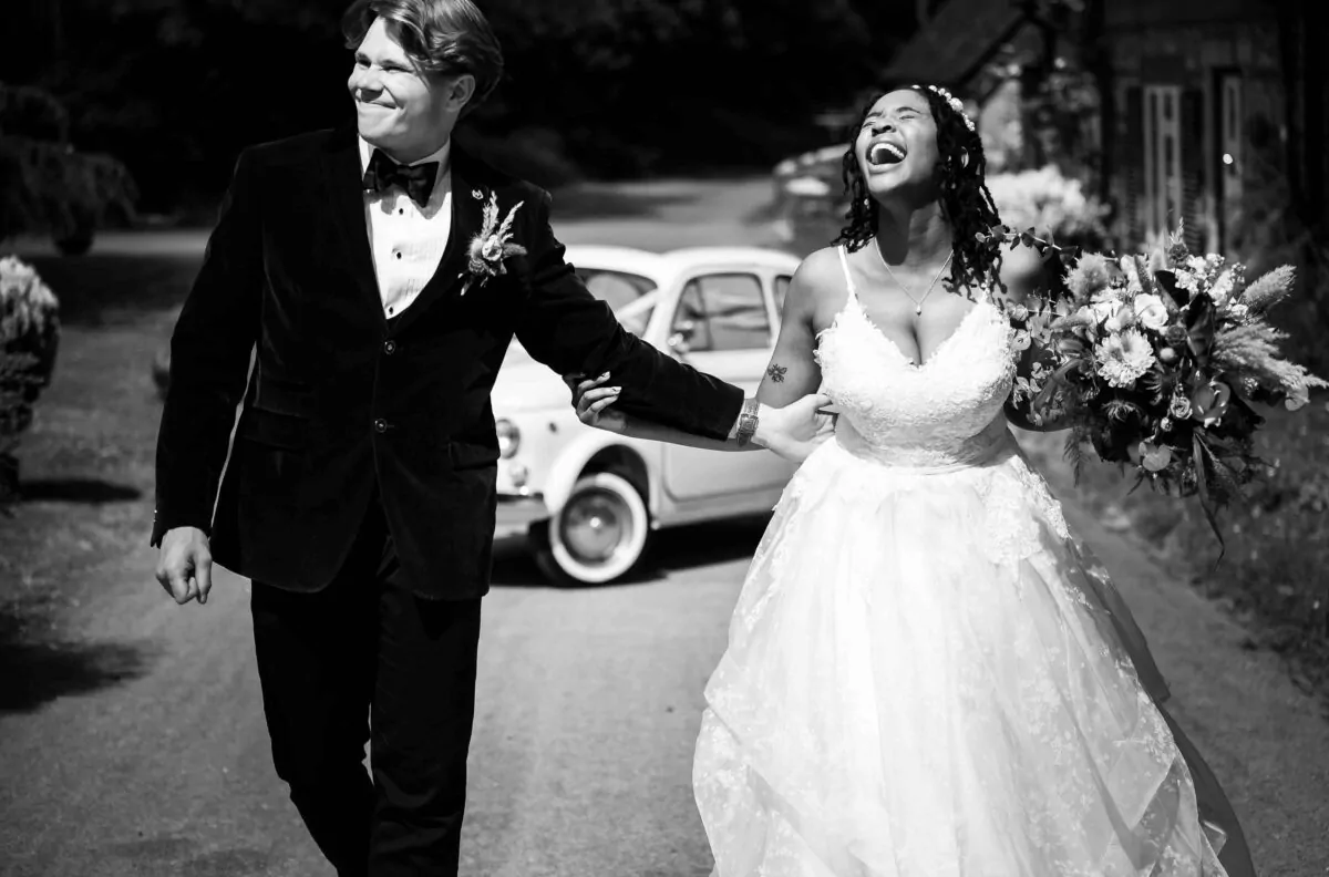 An interracial newlywed couple walks outdoors in bright sunlight, laughing joyfully as they hold hands with a vintage car parked in the background.
