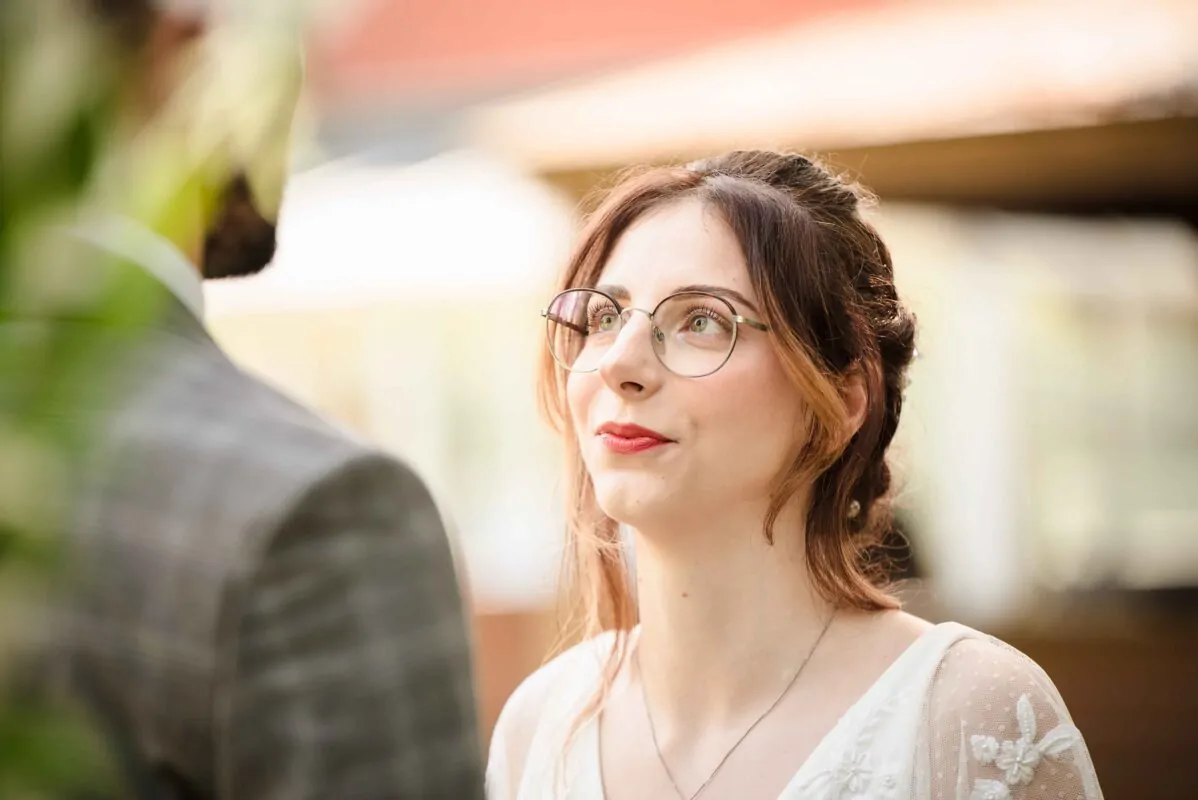 A smiling bride wearing round glasses and a delicate white dress gazes lovingly at her groom during their wedding ceremony.