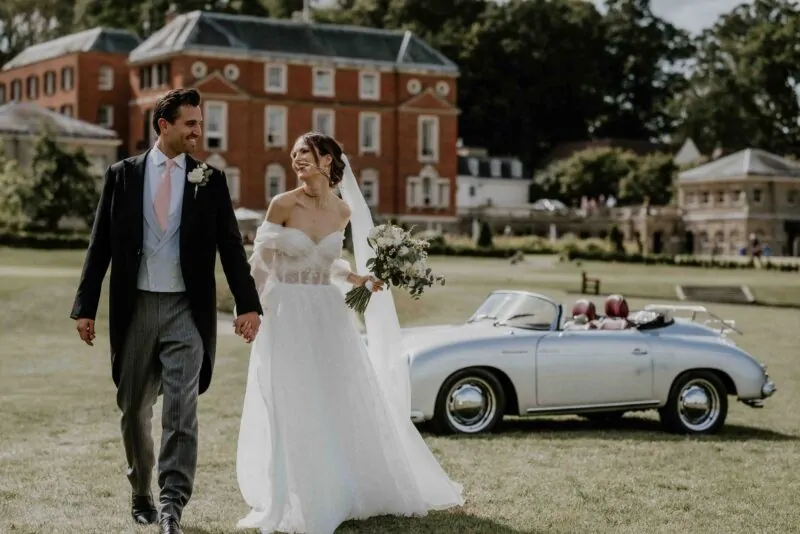 A newlywed couple walk hand-in-hand across the lawn of a grand manor house, passing a silver vintage convertible car while sharing a joyful glance.