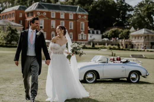 A newlywed couple walk hand-in-hand across the lawn of a grand manor house, passing a silver vintage convertible car while sharing a joyful glance.