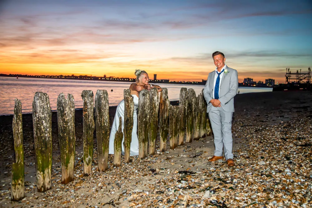 A newlywed couple poses on a pebble beach at dusk, with the bride leaning against wooden groynes and the groom standing nearby against a vibrant sunset sky.