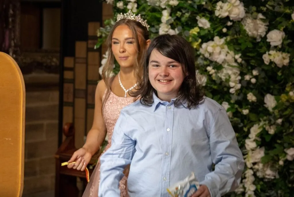 A young smiling boy in a light blue button-down shirt stands in the foreground at a wedding reception near a lush floral backdrop.