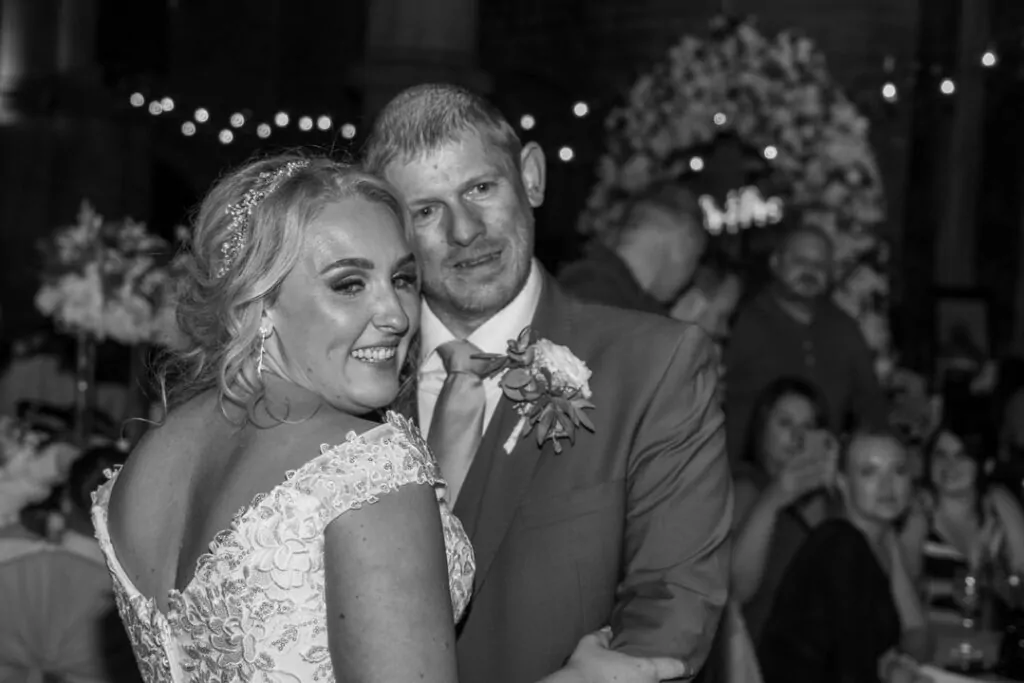 A newlywed couple shares a joyful embrace and smile on the dance floor during their evening reception, captured in black and white.