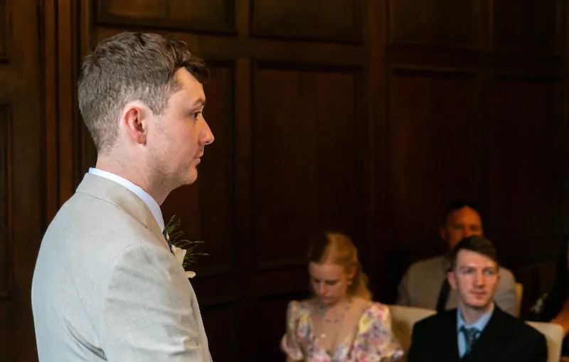 A groom in a grey suit waits at the altar inside a wood-panelled room while guests look on during the ceremony.