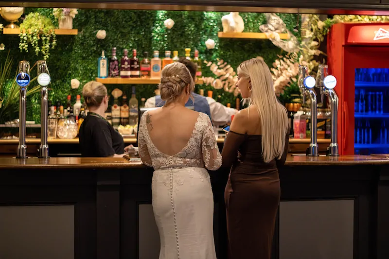 A bride in a lace wedding dress with an open back stands at a pub bar alongside a guest during the drinks reception.