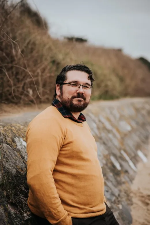 A groom wearing a mustard yellow knit jumper stands outdoors near a stone wall and coastal path on an overcast day.