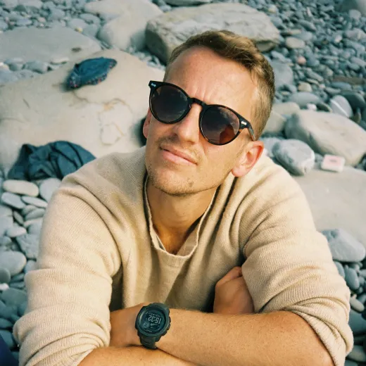 A groom relaxes outdoors on a pebble beach, wearing sunglasses and a casual knit jumper with his arms crossed.