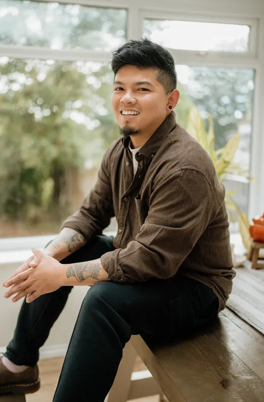 A smiling groom with tattoos wearing a brown shirt sits indoors near a large window, illuminated by soft natural light.